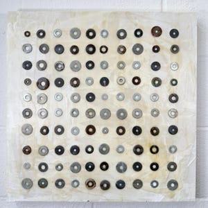 May include: Abstract art piece featuring a grid of metal washers in various sizes and shades of gray, black, and brown. The washers are affixed to a textured, off-white background, creating a visually interesting pattern. The artwork is square in shape.