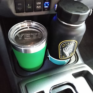 May include: A green Yeti tumbler and a black water bottle with an Orlando, FL, sticker sit in a car's console cup holders. The Yeti tumbler has a silver rim and a clear lid. The water bottle has a black lid.