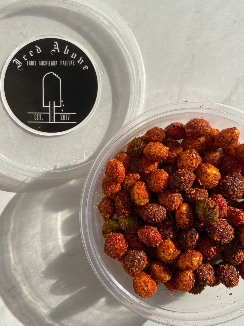 Op de afbeelding: Een doorzichtige plastic container gevuld met rode, oranje en groene snacks bedekt met chili. De container staat op een wit oppervlak. De tekst "Fread Above" is zichtbaar op een zwarte sticker op een tweede doorzichtige plastic container.