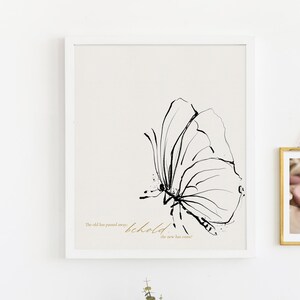 K&ouml;nnte beinhalten: Ein wei&szlig;er gerahmter Druck mit einer Schwarzwei&szlig;-Schmetterlings-Illustration und dem Text "The old has passed away, behold, the new has come". Ein goldfarbener gerahmter Fotoabzug von Babyf&uuml;&szlig;en h&auml;ngt an der Wand &uuml;ber einem goldenen Kerzenhalter.