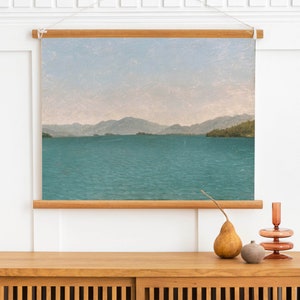 May include: A framed canvas print of a landscape painting depicting a lake with mountains in the distance. The sky is a light blue with a hint of pink. The water is a deep blue-green.