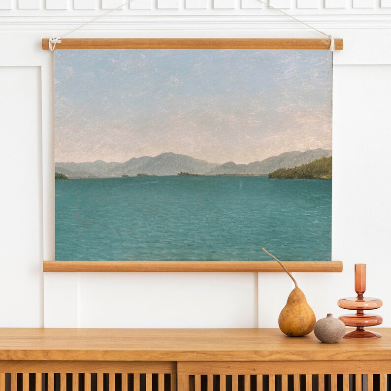 May include: A framed canvas print of a landscape painting depicting a lake with mountains in the distance. The sky is a light blue with a hint of pink. The water is a deep blue-green.