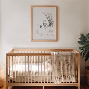 May include: A black and white illustration of Winnie the Pooh and Piglet walking in the woods. The illustration is framed and hanging on a white wall above a wooden cot with a crocheted blanket.