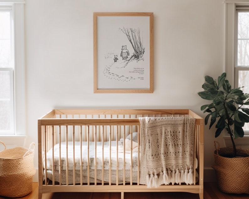 May include: A black and white illustration of Winnie the Pooh and Piglet walking in the woods. The illustration is framed and hanging on a white wall above a wooden cot with a crocheted blanket.