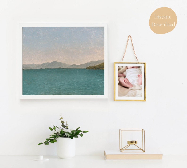 May include: A white frame with a landscape photograph of a lake and mountains. A gold frame with a photograph of a baby's feet. A gold metal stand with a round top and three legs. A white ceramic vase with green leafy plants.