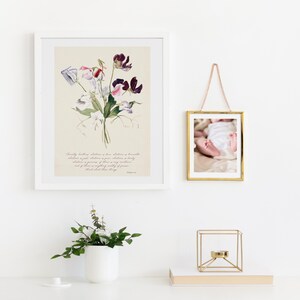 May include: A framed floral print with a white mat and frame, featuring a watercolor illustration of purple, pink, and white sweet pea flowers. A small gold framed photo of a baby's feet hangs nearby. A white vase with greenery and a gold geometric candle holder are on a white surface.
