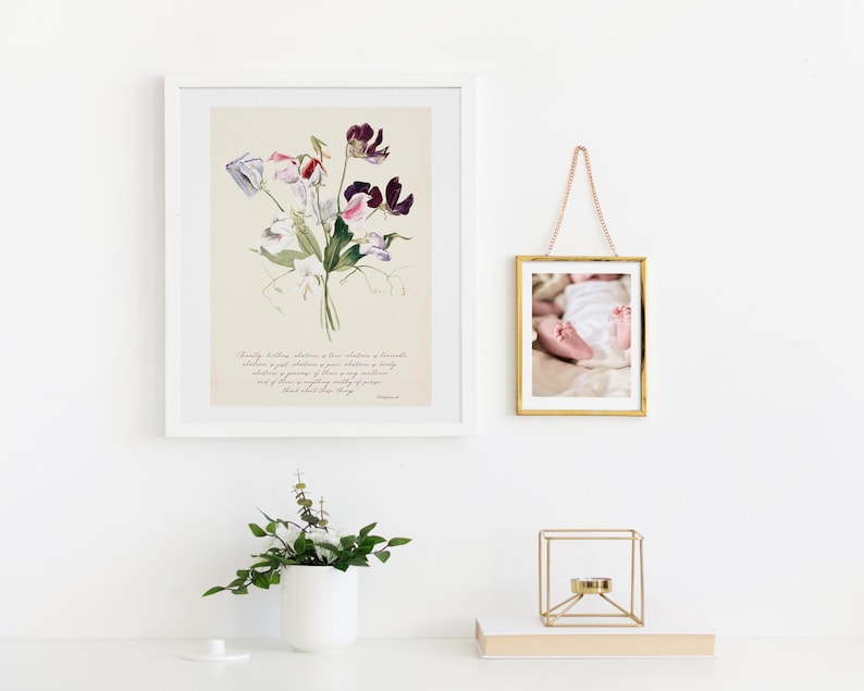 May include: A framed floral print with a white mat and frame, featuring a watercolor illustration of purple, pink, and white sweet pea flowers. A small gold framed photo of a baby's feet hangs nearby. A white vase with greenery and a gold geometric candle holder are on a white surface.
