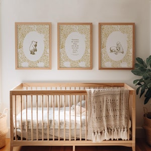 May include: Three framed prints with gold floral patterns and illustrations of Winnie the Pooh characters. The prints are hanging on a white wall above a wooden cot with a white blanket. A potted plant is on the right side of the image.