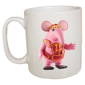 Puede incluir: Taza de cerámica blanca con un ratón de dibujos animados rosa que lleva un disfraz rojo y amarillo. El ratón tiene una nariz larga y orejas grandes. La taza tiene un asa.