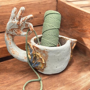 以下が含まれることがあります： 緑色の糸玉を持った手の形をした取っ手が付いたセラミック製の糸巻きボウル。ボウルは白の釉薬で、茶色と青のアクセントが施されています。