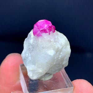 Op de afbeelding: Een helderroze robijnkristal ligt op een witte, getextureerde steen. De edelsteen is gemonteerd op een heldere, rechthoekige acrylbasis, die de natuurlijke schoonheid van het mineraalexemplaar benadrukt. De achtergrond is donkerblauw.