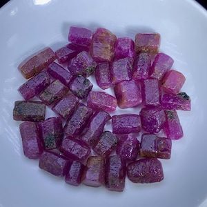 Puede incluir: Una colección de piedras preciosas de rubí en bruto, en varios tonos de rosa y rojo, presentadas en un cuenco blanco. Las piedras en bruto tienen un aspecto natural, sin pulir, que muestra su estructura cristalina. Las piedras preciosas son de diferentes tamaños y formas.