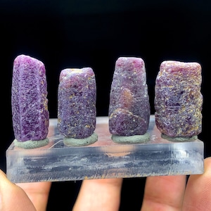 Puede incluir: Cuatro pequeños cristales de piedras preciosas facetados de color púrpura en un soporte de exhibición transparente. Los cristales están dispuestos en una fila, con el cristal más grande en el centro.