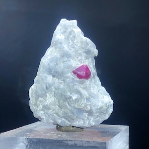 以下が含まれることがあります： 小さなざらざらした灰色の岩に、鮮やかな赤いルビーの結晶が埋め込まれています。岩は透明なアクリル製のディスプレイスタンドの上に置かれています。