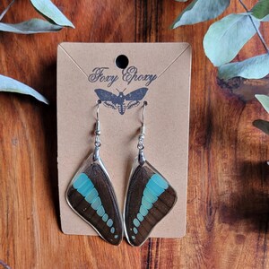 May include: A pair of butterfly wing earrings with a brown and blue design. The earrings are displayed on a brown card with the text "Foxy Epoxy" and a moth illustration. The background is a wooden surface.