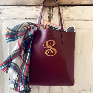 May include: A burgundy leather tote bag with a gold monogram "S" on the front. The bag is hanging on a white wooden surface with a plaid scarf and a magazine inside.