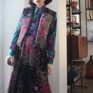 Könnte beinhalten: Ein langer schwarzer Rock mit Blumenmuster und eine grün-rosa gemusterte Weste. Rock und Weste haben ein ähnliches Blumenmuster. Die Person trägt schwarze Schuhe.