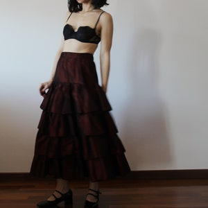 May include: A burgundy tiered maxi skirt with a ruffled hem. The skirt is made of a silky fabric and has a fitted waist.