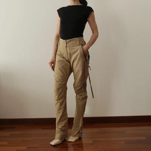 Könnte beinhalten: Ein schwarzes Top mit kurzen Ärmeln und eine beige Cargohose. Die Hose hat mehrere Taschen, Knöpfe und ein einzigartiges Riemendesign. Das Outfit wird durch beige quadratische Ballerinas ergänzt. Der Gesamtstil ist lässig und zweckmäßig.