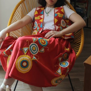 May include: A woman wearing a red skirt with colorful geometric embroidery and a white top with a yellow, blue, and red vest. She is sitting in a yellow wicker chair.