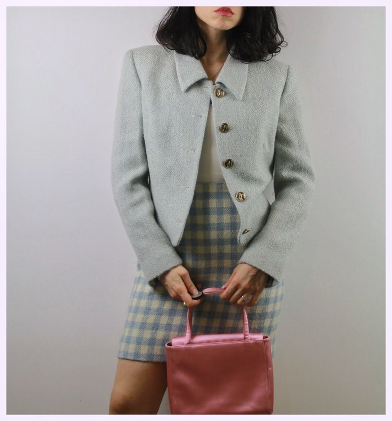 Op de afbeelding: Een lichtblauwe, getailleerde blazer met gouden knopen en een witte kraag. De blazer wordt gedragen over een wit topje en een geruite rok. Een roze handtas wordt in de hand gehouden.