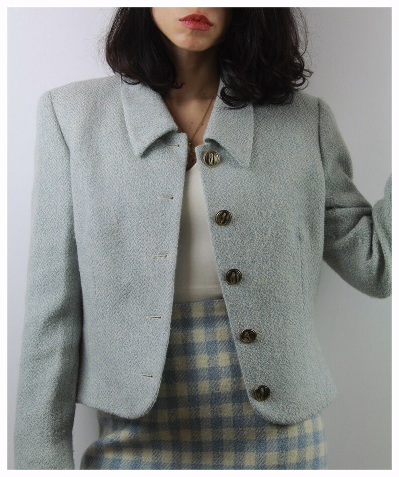 Op de afbeelding: Een lichtblauwe blazer met een herringbone-patroon en gouden knopen. De blazer heeft een getailleerde lengte en een kraag.
