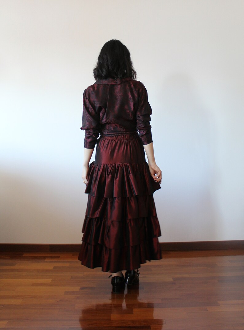 May include: A burgundy brocade blouse with long sleeves and a ruffled skirt. The skirt has multiple layers of ruffles and is a deep burgundy color.