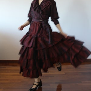May include: A woman wearing a burgundy ruffled skirt and a matching burgundy blouse with a ruffled collar. She is wearing black shoes and is twirling.