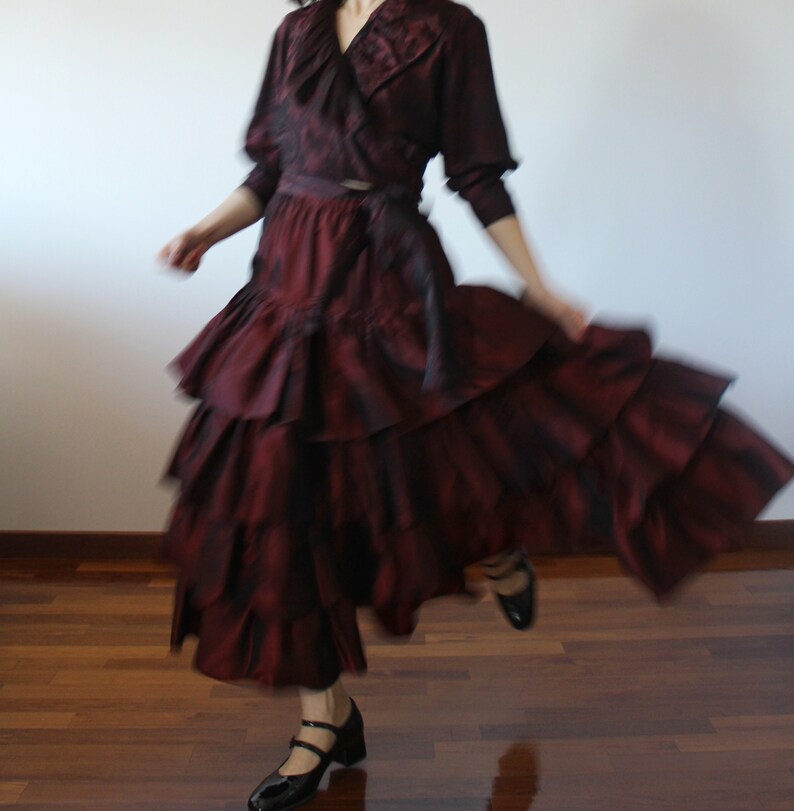 May include: A woman wearing a burgundy ruffled skirt and a matching burgundy blouse with a ruffled collar. She is wearing black shoes and is twirling.