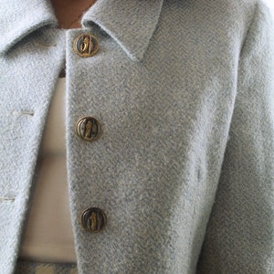 Op de afbeelding: Een lichtblauwe blazer met een herringbone-patroon en gouden knopen. De knopen hebben een decoratief ontwerp.