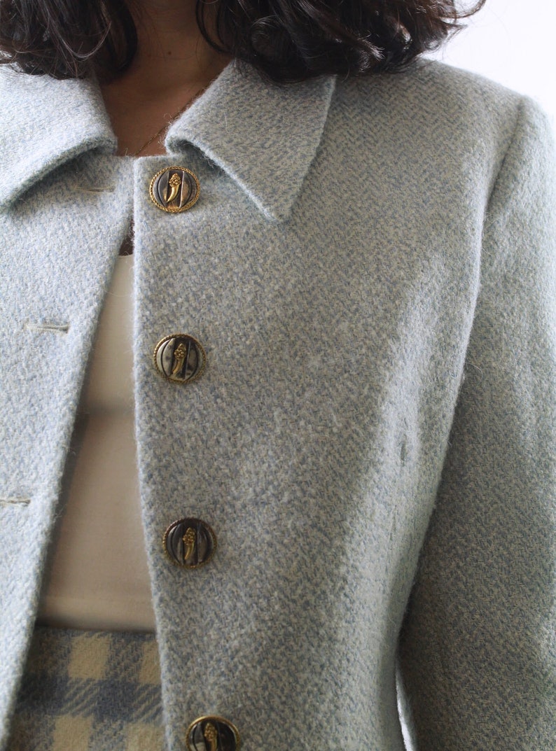 Op de afbeelding: Een lichtblauwe blazer met een herringbone-patroon en gouden knopen. De knopen hebben een decoratief ontwerp.