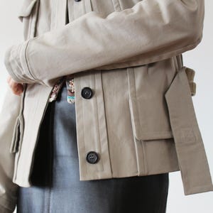 May include: A beige utility jacket with a belt, flap pockets, and button closures. The jacket is worn over a black leather skirt.