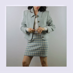 Op de afbeelding: Een lichtblauwe blazer met een wit en lichtblauw geruite rok. De blazer heeft een kraag en gouden knopen.