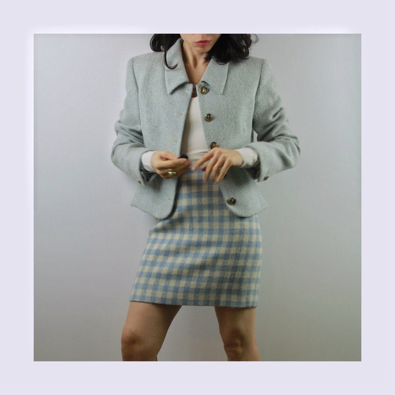 Op de afbeelding: Een lichtblauwe blazer met een wit en lichtblauw geruite rok. De blazer heeft een kraag en gouden knopen.