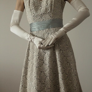 Peut inclure: Une robe vintage en dentelle beige avec un col carré et une ceinture bleu clair. La robe est assortie de longs gants blancs en satin. Un collier de perles ajoute une touche d'élégance. La robe présente un motif floral en dentelle.
