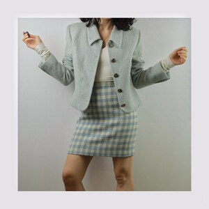 Op de afbeelding: Een lichtblauwe blazer en bijpassende rokset. De blazer heeft een kraag en knoopsluiting. De rok is een geruit patroon met witte en lichtblauwe vierkanten.