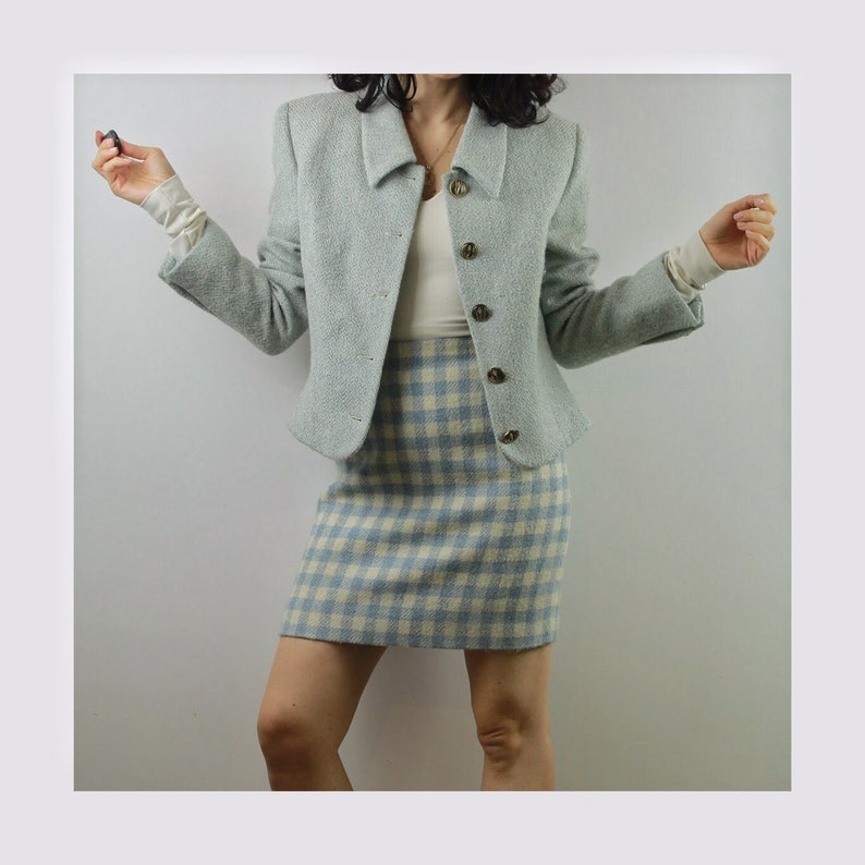Op de afbeelding: Een lichtblauwe blazer en bijpassende rokset. De blazer heeft een kraag en knoopsluiting. De rok is een geruit patroon met witte en lichtblauwe vierkanten.