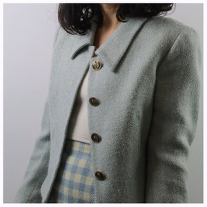 Op de afbeelding: Een lichtblauwe blazer met een wit en lichtblauw geruite rok. De blazer heeft gouden knopen met een zwart ontwerp.