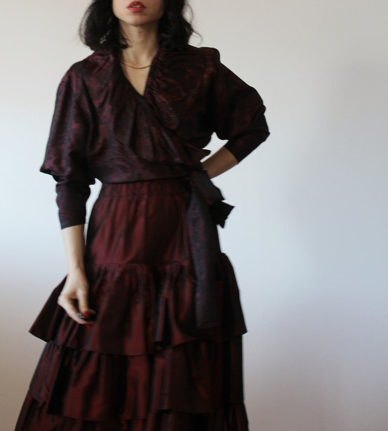 May include: A burgundy silk dress with a ruffled skirt and a wrap top. The top has a V-neckline and a long sleeve.