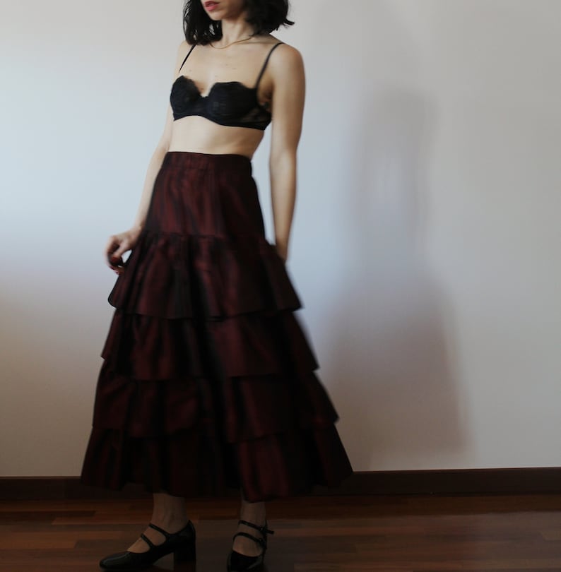 May include: A burgundy tiered maxi skirt with a ruffled hem. The skirt is made of a silky fabric and has a fitted waist.