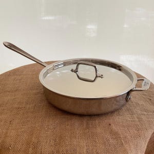 May include: Stainless steel skillet with a lid. The skillet has a long handle and a smaller handle on the side. The lid has a square handle. The skillet is sitting on a brown surface.