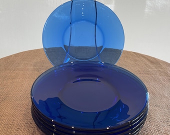 May include: A stack of six cobalt blue glass plates with a clear glass plate holder. The plates are round with a slightly raised edge. The plates are displayed on a brown surface.