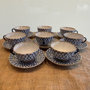 May include: A set of eight porcelain teacups and saucers. The cups and saucers have a white interior and a blue and white lattice pattern on the exterior. The rims are trimmed with gold, and the handles are dark blue.