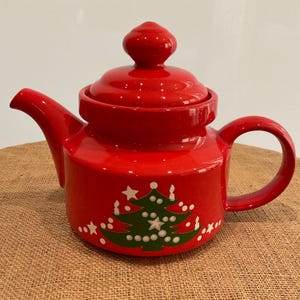 May include: A red ceramic teapot with a lid and handle. The teapot features a Christmas tree design with white stars and green accents. The teapot is designed for brewing tea.