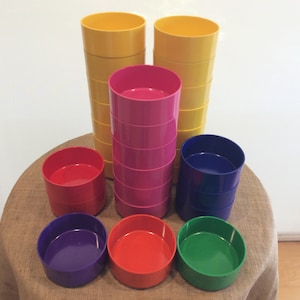 May include: A set of colorful plastic stacking bowls in yellow, pink, red, blue, and green. The bowls are stacked in columns on a brown burlap surface.