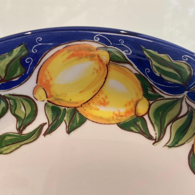 Puede incluir: Plato de cer&aacute;mica con un dise&ntilde;o pintado a mano que presenta dos limones amarillos con reflejos anaranjados y hojas verdes. El borde es azul con remolinos decorativos blancos. El plato tiene un interior blanco.