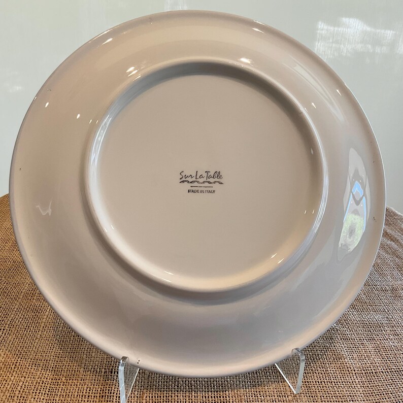 Puede incluir: Un plato de cer&aacute;mica blanca con un borde ligeramente elevado. La parte inferior presenta el texto "Sur La Table" en escritura negra, con "MADE IN ITALY" debajo. El plato se exhibe sobre un soporte transparente, mostrando su dise&ntilde;o.