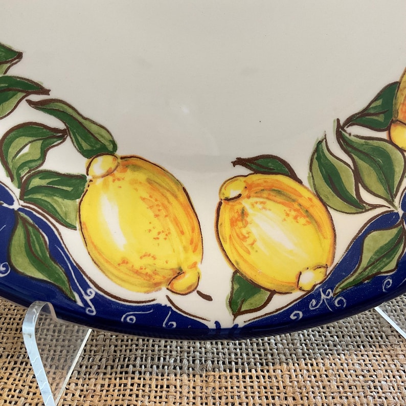 Puede incluir: Un plato de cer&aacute;mica blanco con borde azul, decorado con limones amarillos y hojas verdes pintados a mano. El plato tiene un dise&ntilde;o detallado, perfecto para servir alimentos o como pieza decorativa.