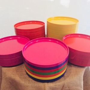 May include: A stack of bright red, yellow, and pink plastic plates. The plates are stacked in a pyramid shape on a brown burlap surface.