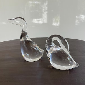 May include: Two clear glass goose figurines. One goose has its head up, and the other has its head curved down. The glass is transparent, allowing light to pass through. The figurines are on a dark brown surface.